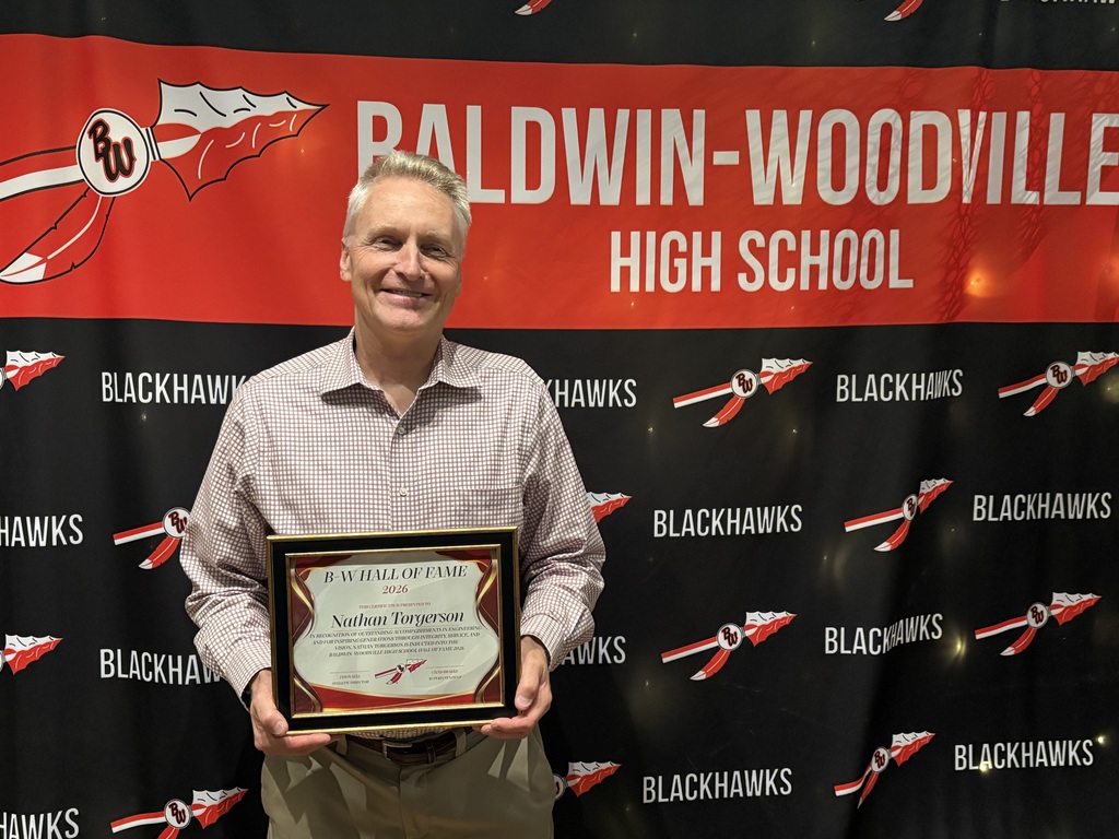 Nathan Torgerson standing in front of a red and black Baldwin-Woodville Blackhawks photo backdrop, smiling and holding his framed 2026 Hall of Fame induction plaque.