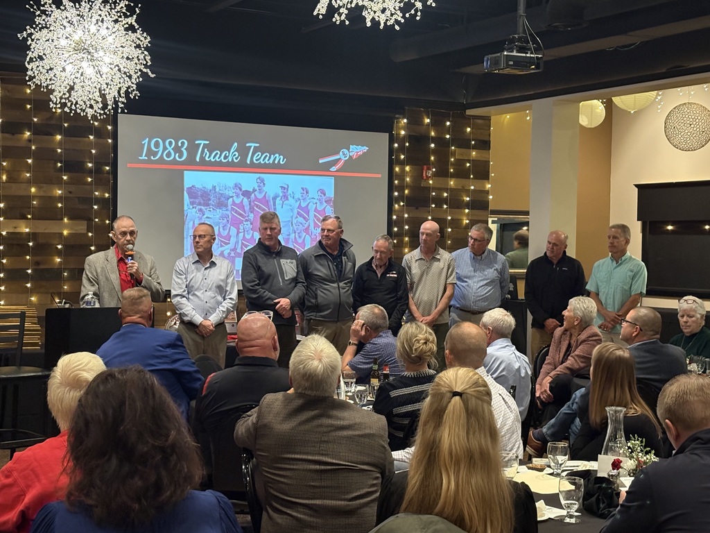 A speaker stands at a podium addressing the audience inside a banquet venue while members and coaches of the 1983 Track Team stand on stage in front of a projection screen displaying their team photo.