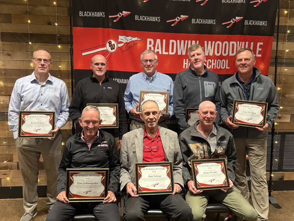 Members and coaches of the 1983 Boys Track Team posed together in front of a red and black Baldwin-Woodville Blackhawks backdrop, holding their individual framed 2026 Hall of Fame induction plaques.