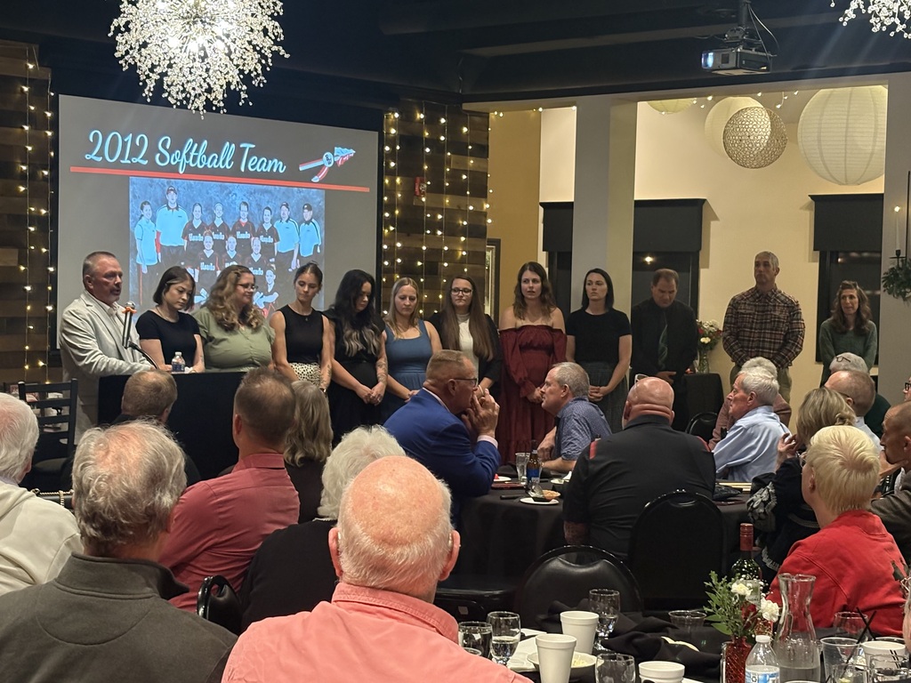 A speaker at a podium addresses an audience seated at banquet tables, with members and coaches of the 2012 Softball Team standing on stage in front of a projection screen showing their team photo.