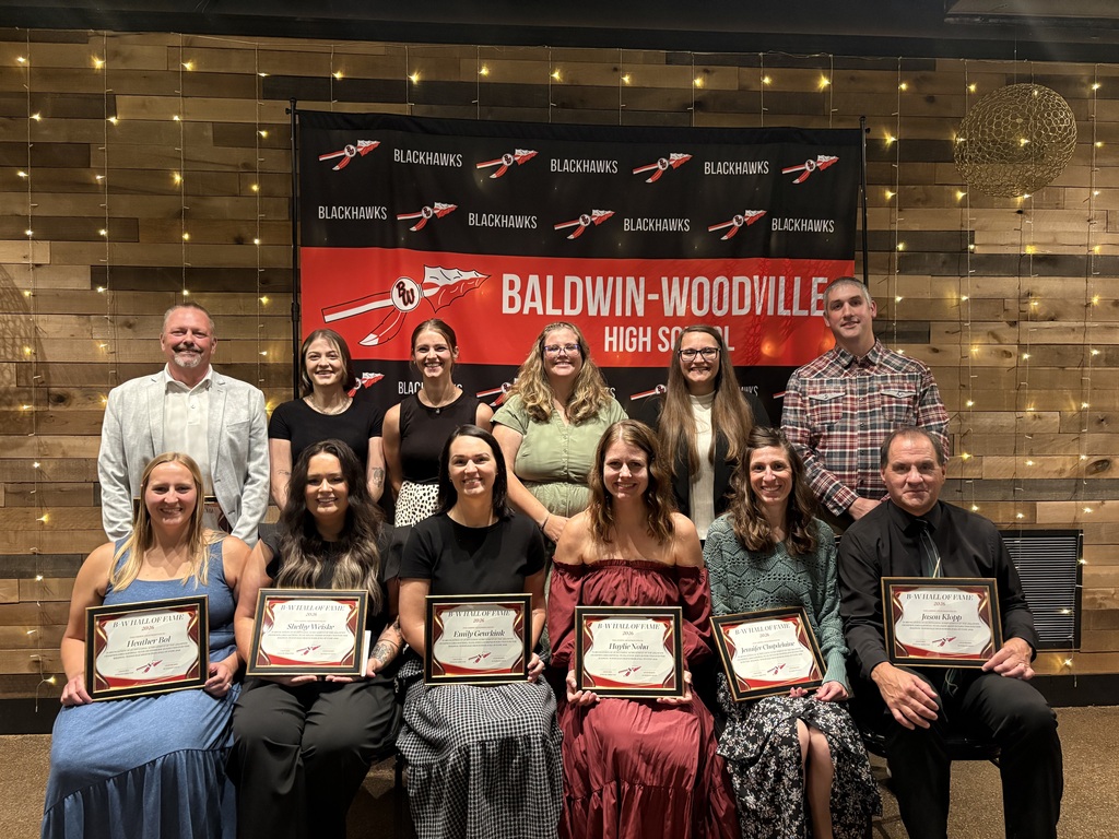 Members and coaches of the 2012 Girls Softball Team smiling together in front of a red and black Baldwin-Woodville Blackhawks backdrop while holding their framed 2026 Hall of Fame induction plaques.