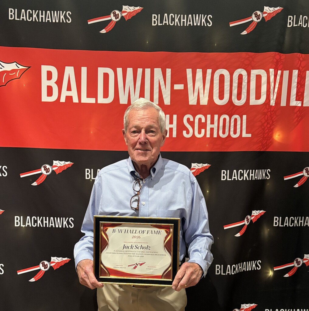 Jack Scholz standing in front of a red, black, and white "Baldwin-Woodville High School Blackhawks" step-and-repeat backdrop. He is wearing a blue button-down shirt and khakis, proudly holding his framed "B-W Hall of Fame 2026" induction plaque.