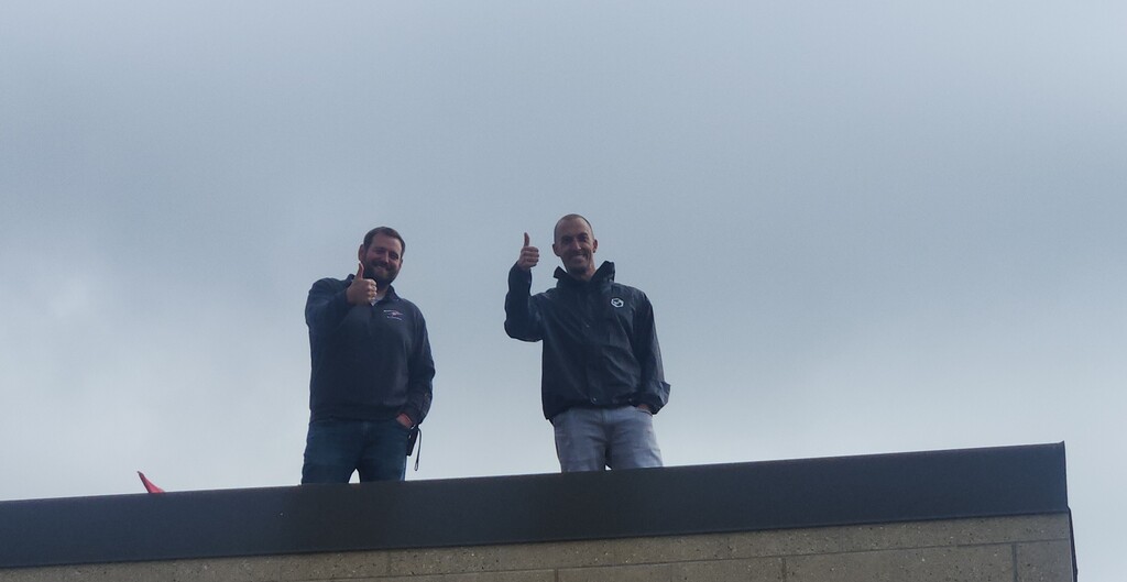 "Two school leaders, Mr. Sbertoli and Mr. Vande Berg, standing on the flat roof of Greenfield Elementary under an overcast sky, smiling and giving thumbs-up gestures to celebrate a successful fundraiser."