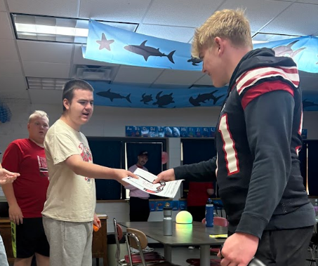 A student in a cream-colored t-shirt presents a hand-colored paper jersey to a smiling high school football player wearing a black uniform. Another student is watching the interaction from behind. A 'shark' themed banner is hanging in the background, contrasting with the focus on district connections.