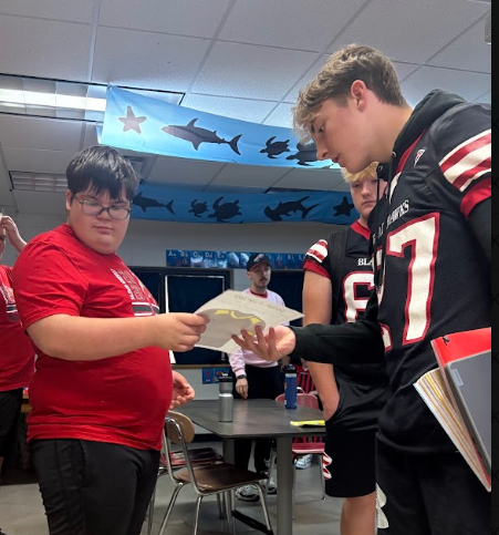 A medium shot of a high school student athlete in a black number 87 football uniform accepting a hand-colored paper football jersey from another student. The student in glasses is holding the paper jersey out, creating a direct connection. A district staff member is visible in the background.