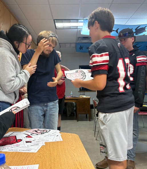 A high school football player in a black number 13 jersey holds up a completed hand-colored paper football jersey to show a staff member and other students. The group is gathered around a table piled with paper jerseys, illustrating the scale of the student engagement activity.