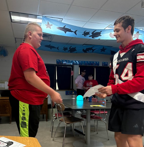 Two high school students inside a classroom. One student in a red shirt receives a hand-colored paper football jersey from a student athlete wearing a black and red football uniform with the number 54.