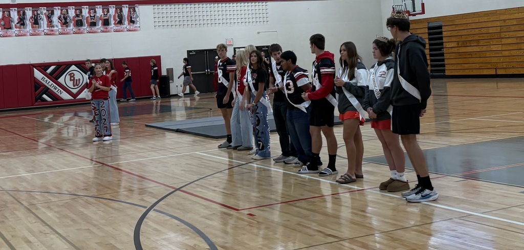 Baldwin-Woodville Homecoming court members wearing royalty sashes and crowns standing in a line along the gymnasium floor facing the crowd.