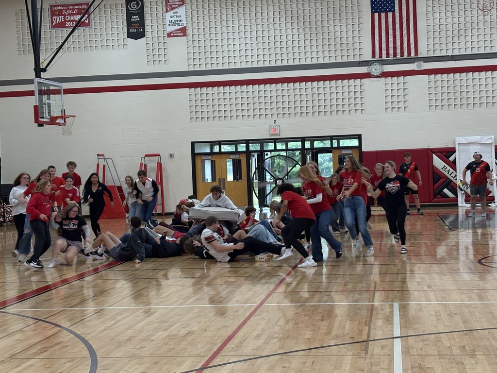 High school students participating in an energetic pep fest game, running across the gymnasium floor while a group tumbles onto a mat in the center.