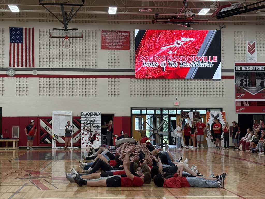 High school students lying side-by-side in a long row across the gym floor as a group tunnel during a pep fest game, with a large video display in the background reading "Baldwin-Woodville Home of the Blackhawks."