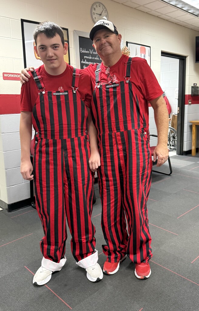 A young man and an older man standing side-by-side inside a school hallway, both smiling and wearing matching red-and-black striped game day bib overalls over red t-shirts to show school spirit.