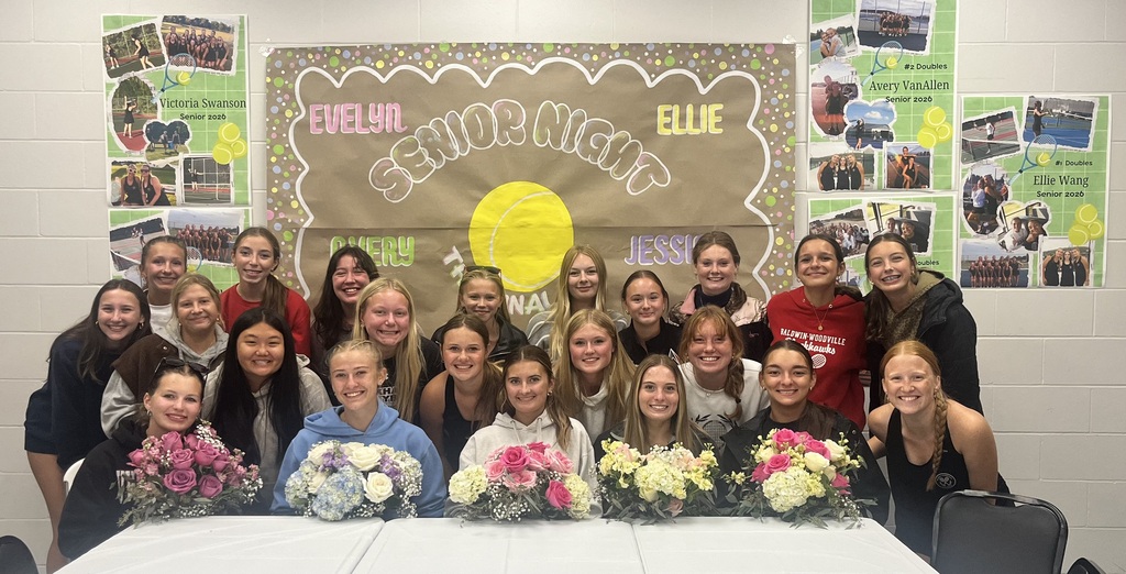 Group photo of high school girls' tennis team celebrating Senior Night. Five seniors sit at a front table holding colorful floral bouquets, flanked by their teammates, with a handmade "Senior Night" banner and photo posters on the wall behind them.