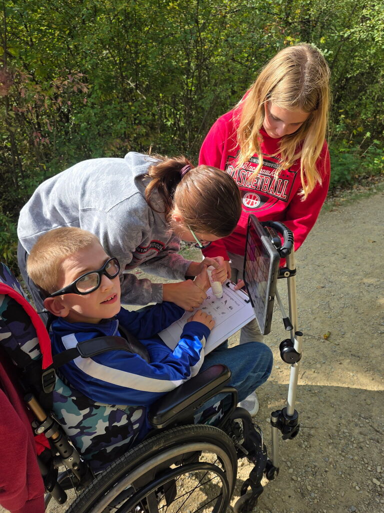 Three students collaborating outdoors on a nature scavenger hunt worksheet clipped to a clipboard, with one student sitting in a wheelchair while two classmates lean in to help.