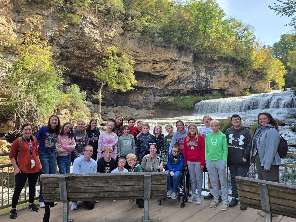 A large group of middle school students and staff posing together on a wooden viewing platform in front of a wide, cascading waterfall and rocky cliff backdrop.