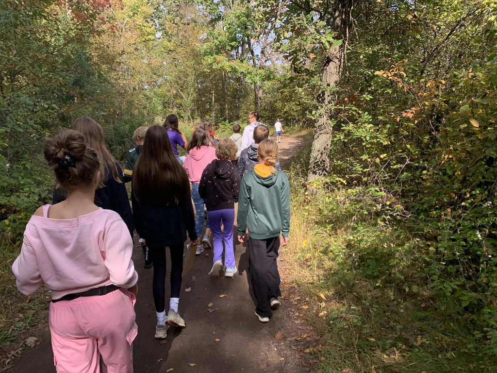 A line of students hiking away down a dirt trail wrapped in lush, green forest foliage on a sunny autumn day.