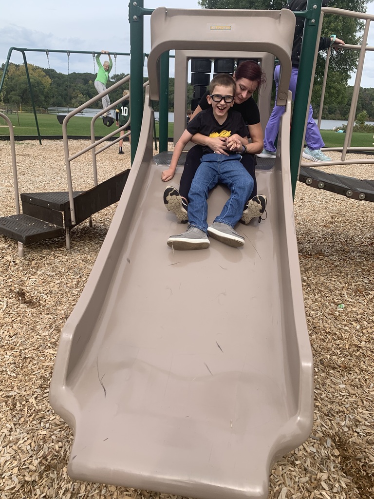 A smiling student and an adult companion riding down a wide playground slide together on a sunny day.