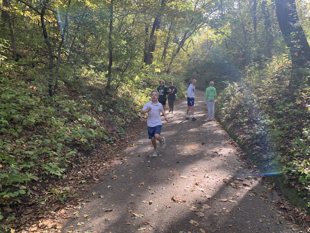 A student jogging cheerfully down a shaded asphalt park path surrounded by woods, with classmates walking behind him.