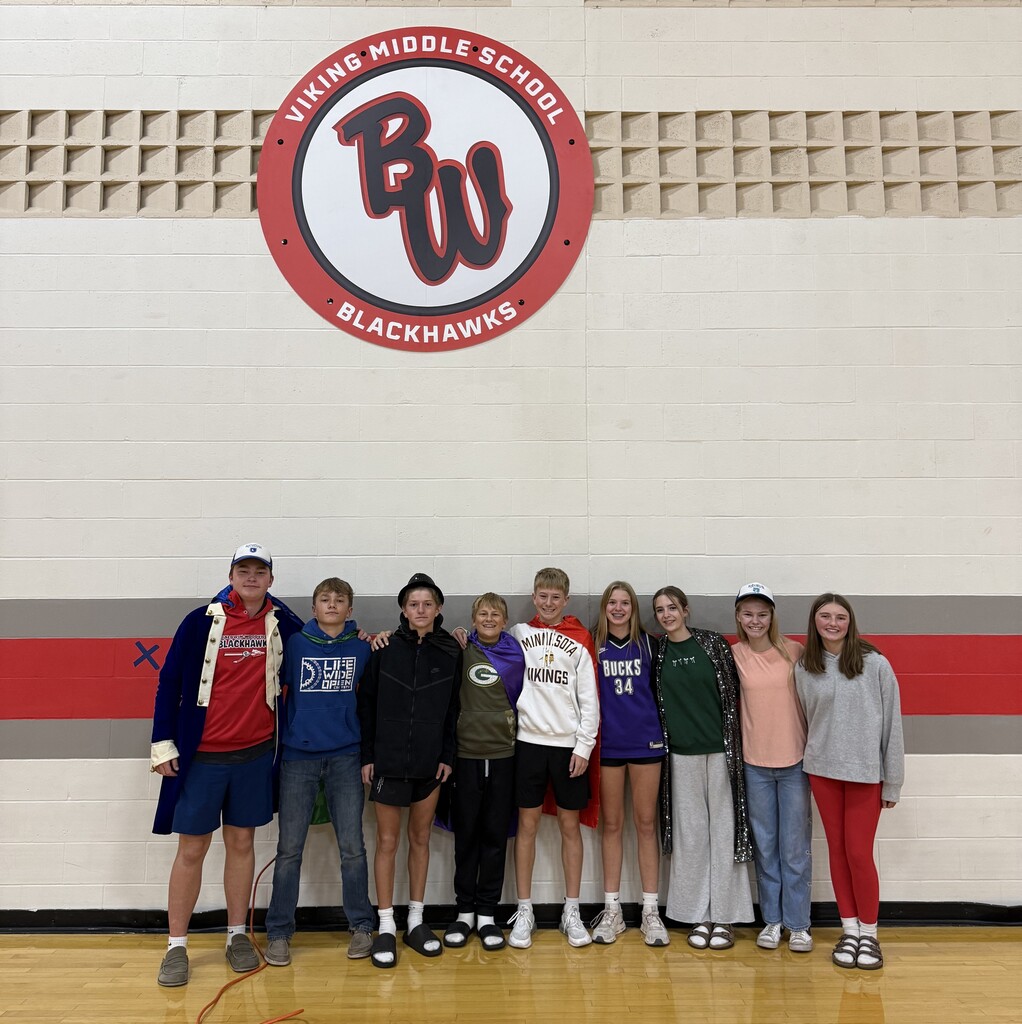 A group of Viking Middle School RCA House Leaders standing in a gym underneath the round "Viking Middle School Blackhawks" wall sign. Five students in the center stand with their backs to the camera, wearing colorful capes showing their RCA House names—Rêveur, Isibindi, Altruismo, Nukumori, and Amistad. Four student leaders stand on either end pointing inward toward the group in capes.