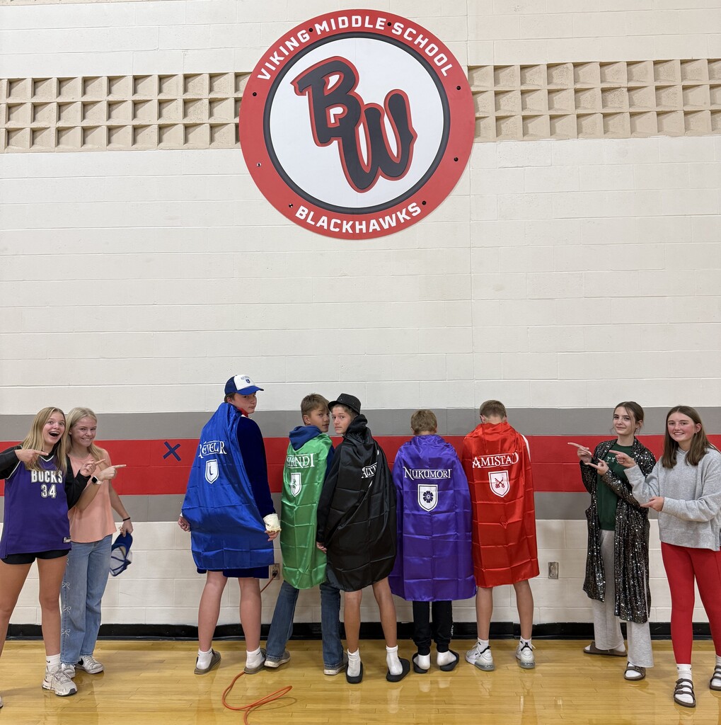 A group of nine smiling Viking Middle School RCA House Leaders standing together in a gym in front of the "Viking Middle School Blackhawks" wall sign, dressed in fun spirit gear, jerseys, hats, and colorful capes.