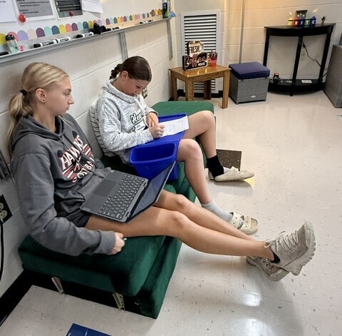 Two middle school students lounge comfortably on a green sofa in a classroom, utilizing flexible seating to work together on their laptops and notebooks.