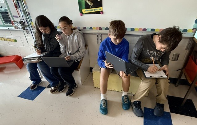 Four middle school students sitting side-by-side on low classroom benches, focusing intently on their clipboards, notebooks, and laptops.