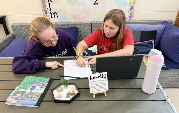 wo middle school students sitting together at a wooden patio-style table in a classroom, collaborating on a fall writing assignment using a laptop and notebook.