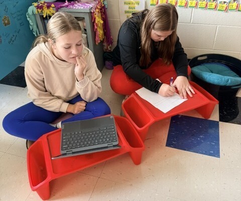 Top-down view of two students sitting cross-legged on the floor, collaborating using red portable lap desks with paper and a laptop.