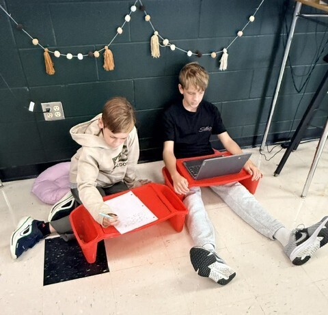 Two students sitting on the classroom floor using red lap desks to work on writing and research tasks on a laptop and paper.