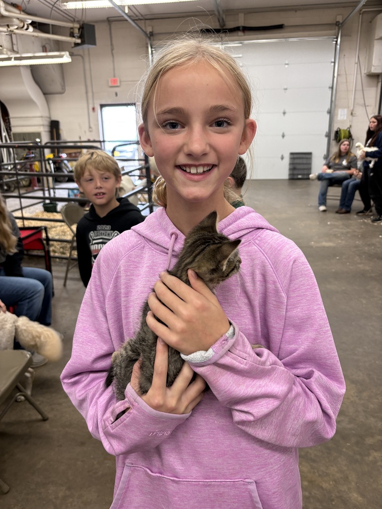 A smiling young girl in a pink hoodie holding a small tabby kitten inside the high school shop area during a petting zoo event.