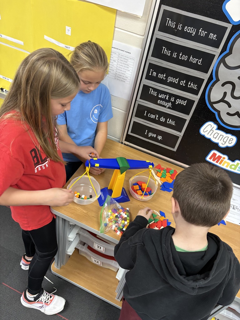 Three students carefully adding colorful plastic pieces to a yellow and blue pan balance scale next to a classroom "Growth Mindset" display.