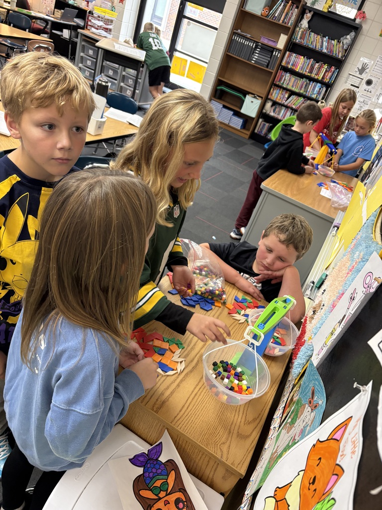 A group of students gathered around a table using a balance scale to weigh colorful plastic cubes and geometric shapes.