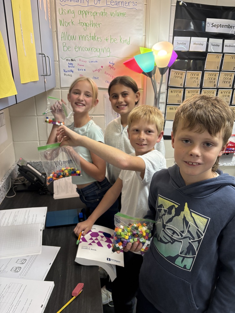 Four smiling 4th-grade students holding up plastic bags filled with colorful plastic measuring cubes in a classroom.