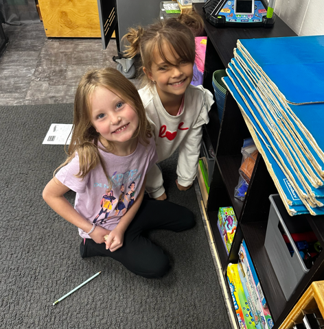 Two young girls smiling at the camera while sitting on the classroom rug next to a green pencil and activity sheet