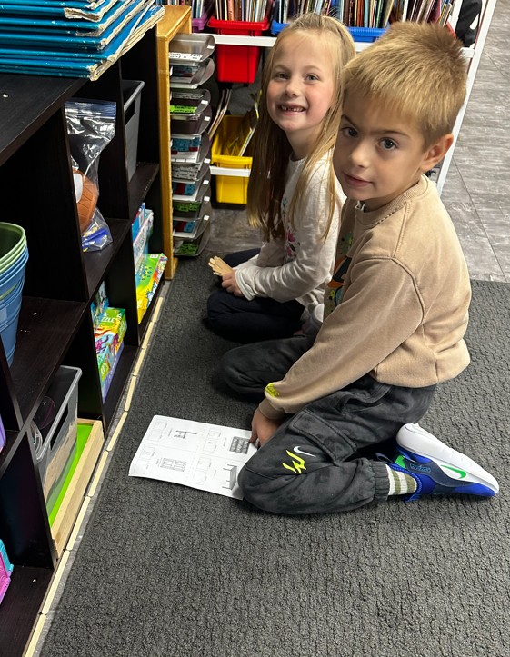 A young boy and girl sitting on the floor next to a line of popsicle sticks being used as a measuring tool.