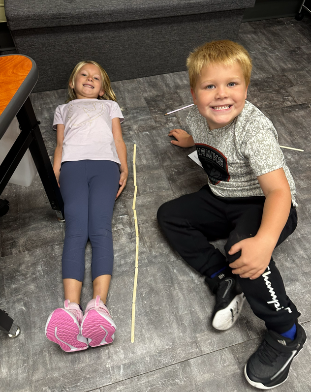 A young boy looking up at the camera while using popsicle sticks to measure a student lying down