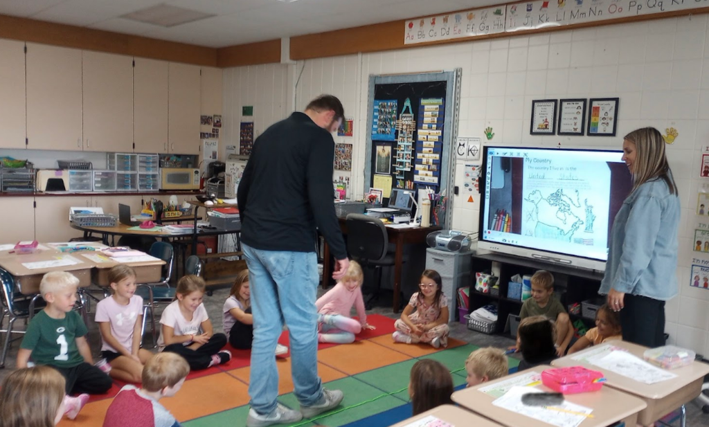 An adult male teacher walking along a green line taped to the rug while kindergarten students and another teacher watch him count his footsteps.