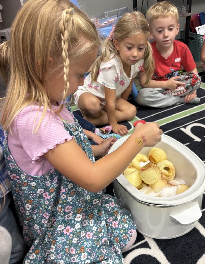 A young girl in a floral dress carefully places ingredients into a slow cooker filled with peeled, sliced apples while two classmates watch attentively.