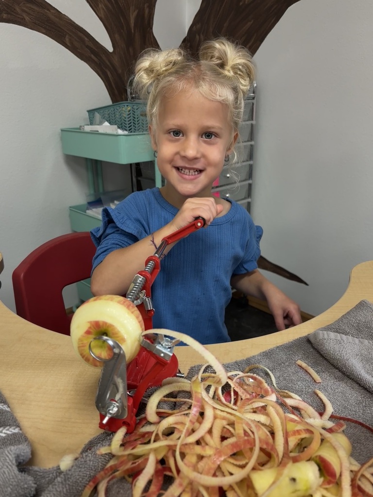 A smiling young girl with pigtail buns uses a red apple peeler at a table covered in fresh apple peels
