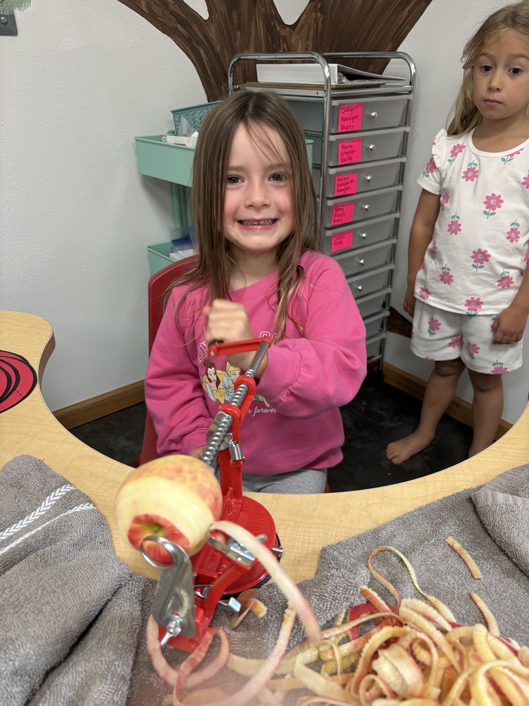 A smiling young girl in a pink sweater turns the handle of a red manual apple peeler at a classroom table.