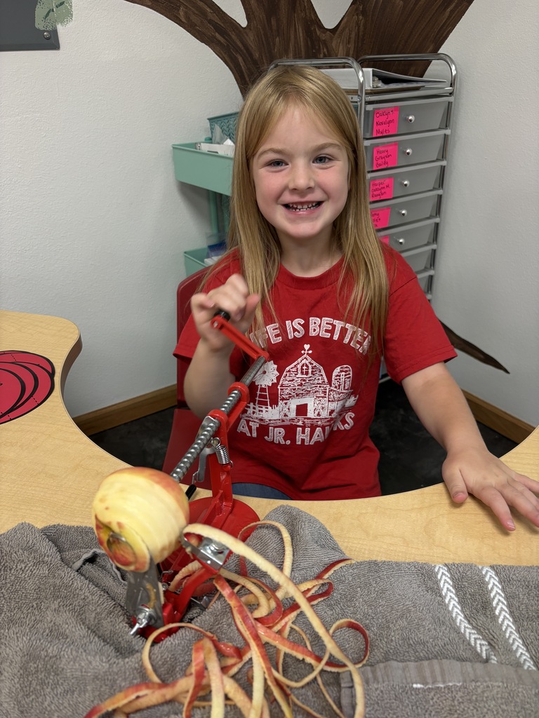 A smiling young girl in a red shirt operates a hand-cranked apple peeler, surrounded by long spiral apple peels.