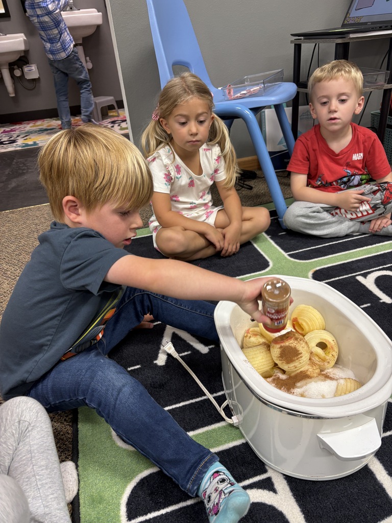 A young boy in a blue shirt shakes cinnamon over peeled apples in a slow cooker, with two classmates sitting nearby on the classroom rug watching