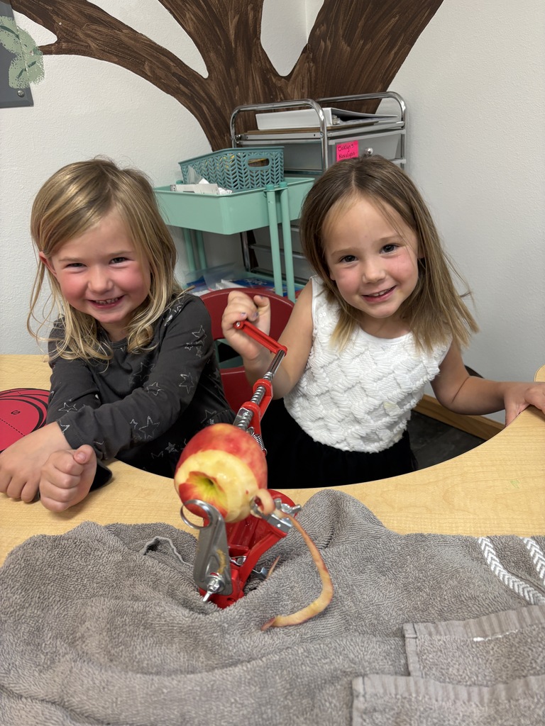 Two young girls smile at the camera while taking turns operating a red apple peeler.