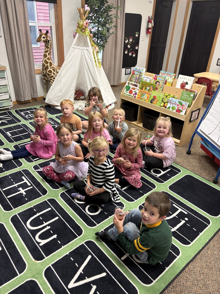 A group of 4K students sit together on the classroom carpet, smiling and enjoying small cups of fresh homemade applesauce with spoons.
