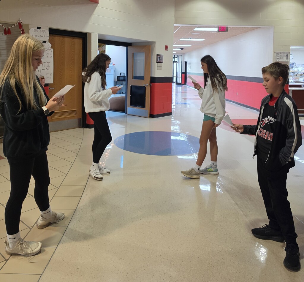 Four middle school students stand in a school hallway in two pairs, holding papers and conversing with each other in Spanish.