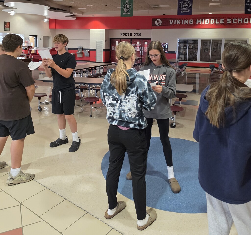 Viking Middle School students stand in pairs in the commons area near the gym, holding practice worksheets and conversing in Spanish