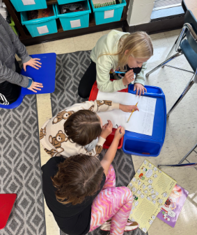High-angle view of three 4th graders sitting on the floor around a small tray table, working together with pencils and markers to create their main idea "graffiti" poster.