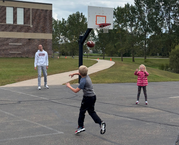A student throws a football toward a basketball hoop on the playground while a principal and classmate look on.