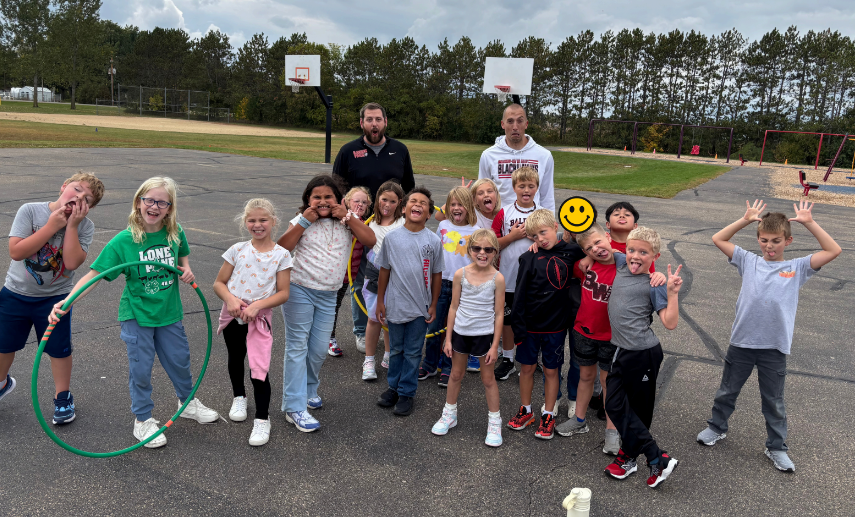 A group of smiling elementary students poses outside on the playground alongside their school principals, holding hula hoops and making fun faces for the camera.