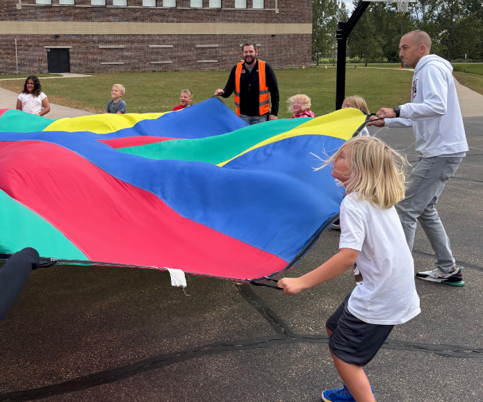 Elementary students and school principals lift a large, colorful play parachute together outdoors on the blacktop