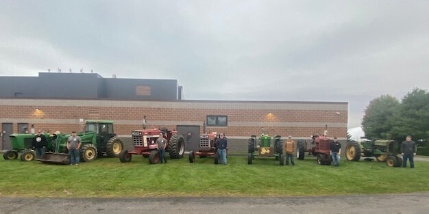 Here are two options for the image alt text, depending on where you are using it: Detailed Alt Text (Best for Web Accessibility): Six high school FFA members standing outdoors in front of a school building on a grassy lawn, each posed next to a distinct vintage or modern tractor lined up side-by-side. From left to right, the row includes green John Deere tractors and red International Harvester tractors.